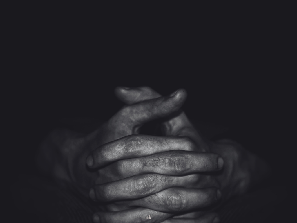 A black and white image of folded hands with blackness all around them.